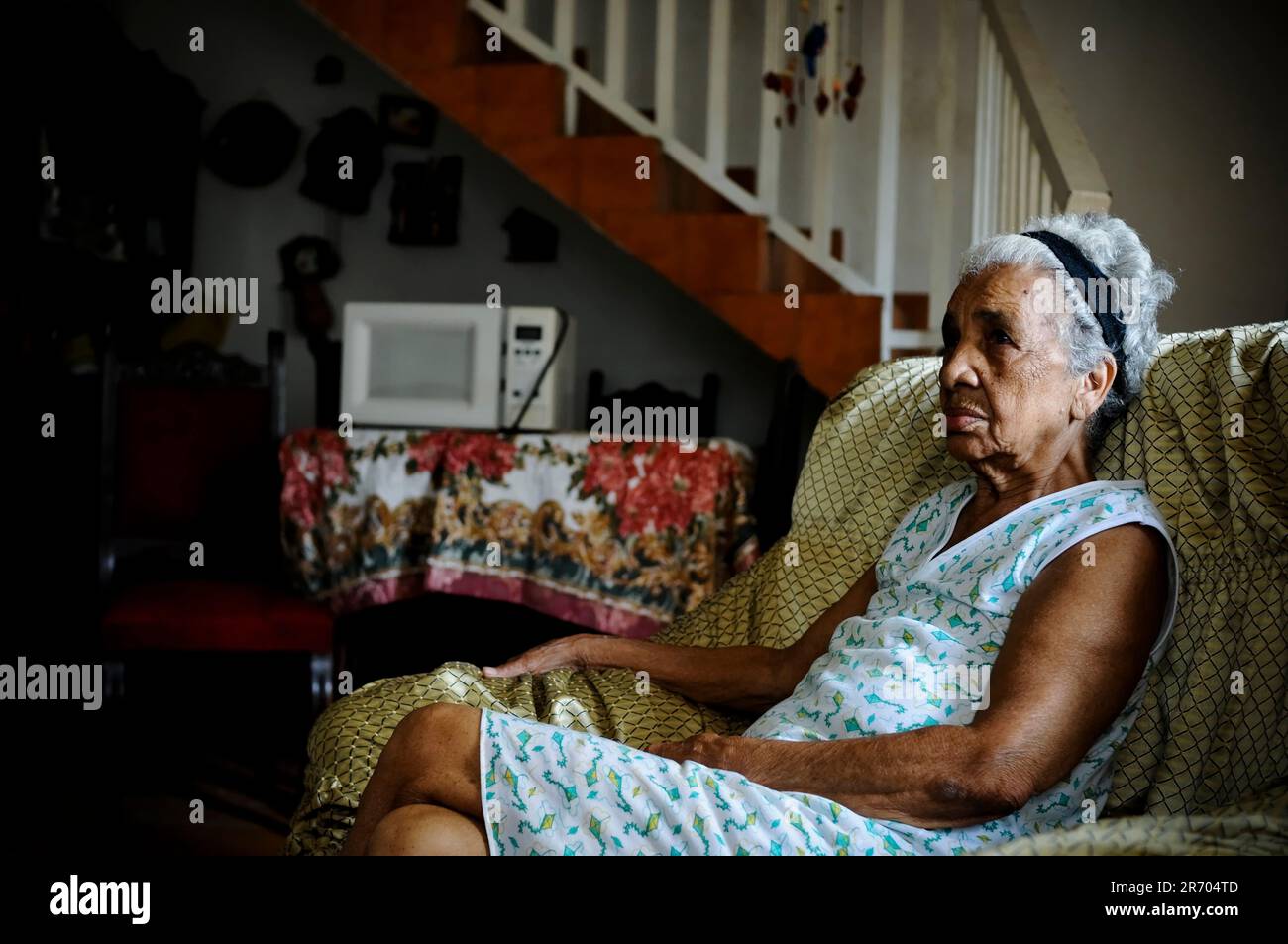 An elderly woman affected by water and electricity rationing sits somberly in her living room in a slum in Caracas, Venezuela. Stock Photo