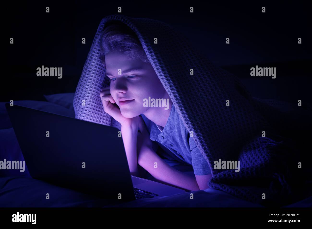 addiction. Teenage boy using laptop under blanket on bed at