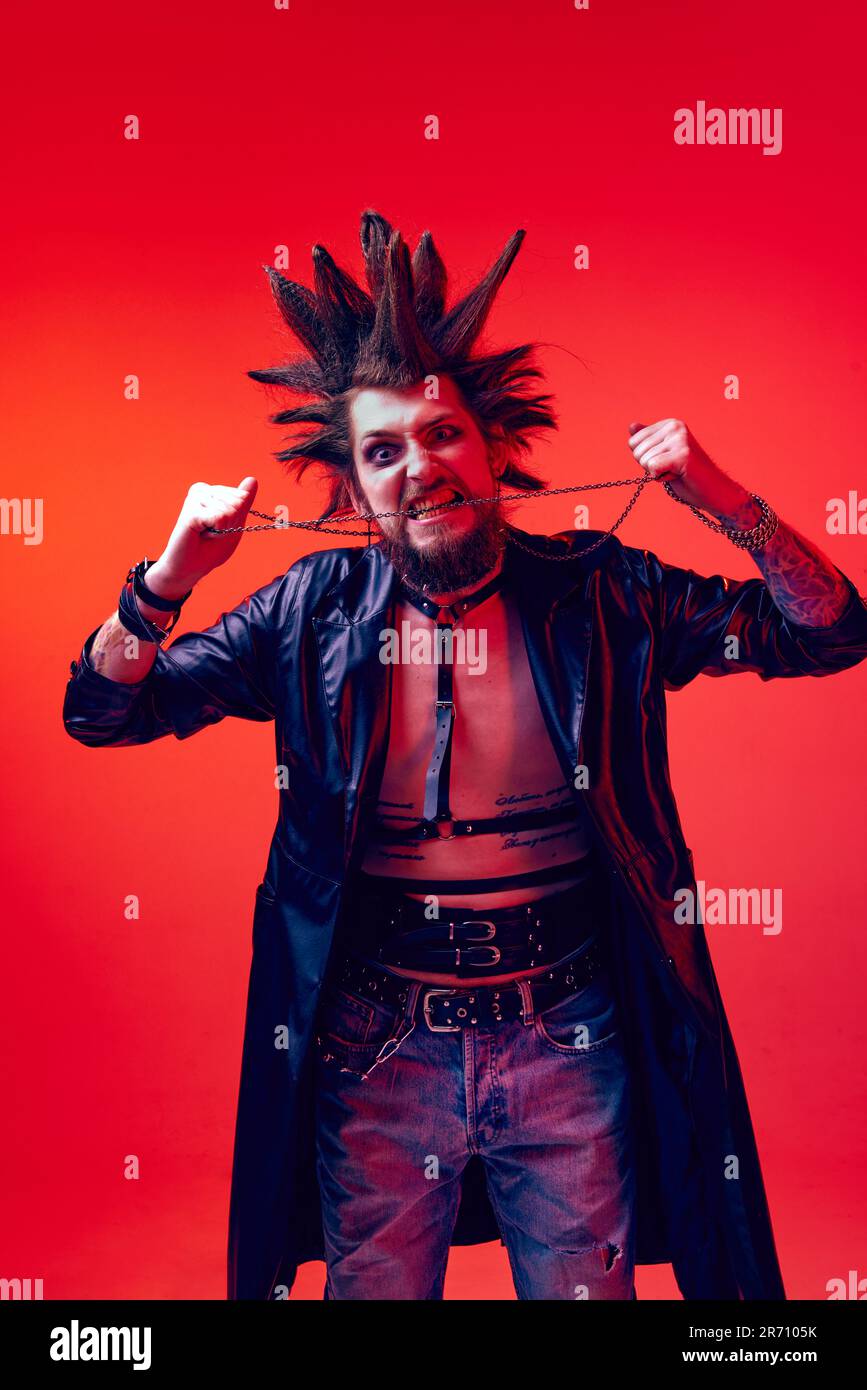 Angry and crazy look. Portrait of young man, punk with crazy hairdo and makeup posing against red studio background in neon light Stock Photo