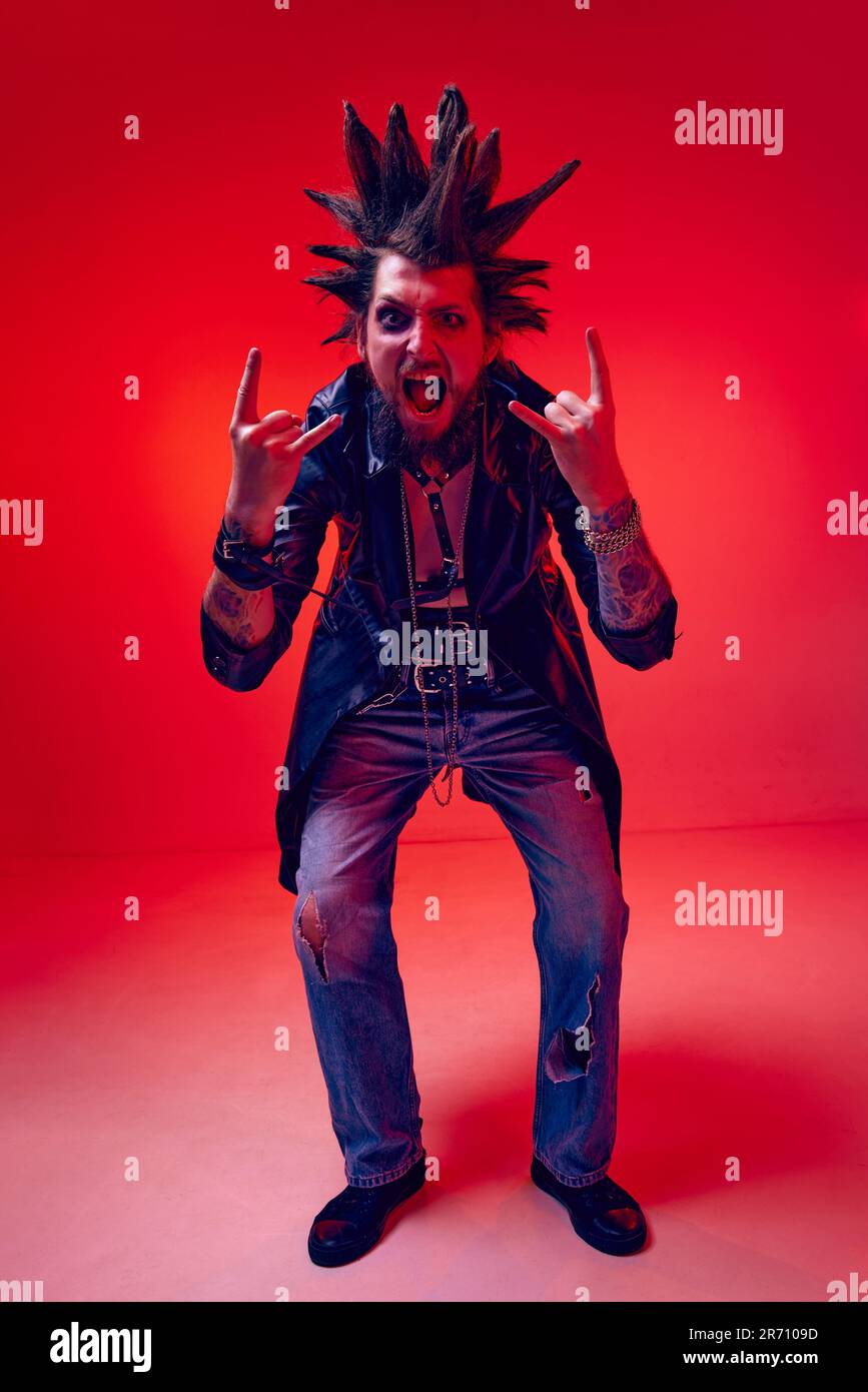 Rock and roll. Emotive, expressive young man, punk with makeup and strange hairdo posing against red studio background in neon light Stock Photo