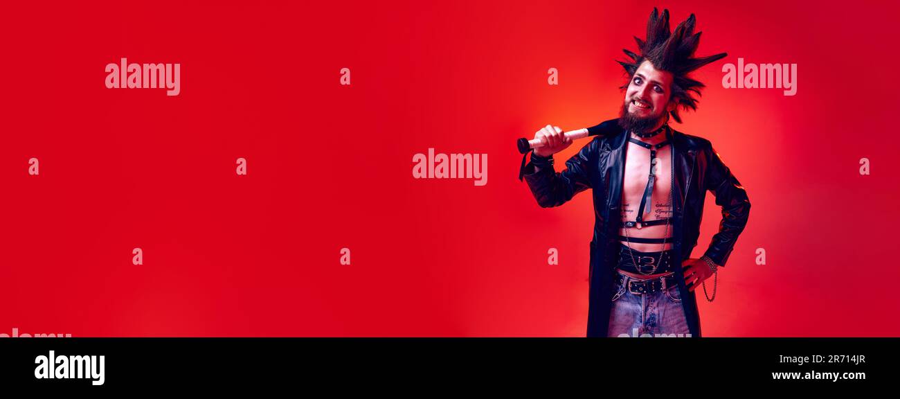 Crazy looking emotive young man, punk with expressive hairdo posing with bat against red studio background in neon light Stock Photo