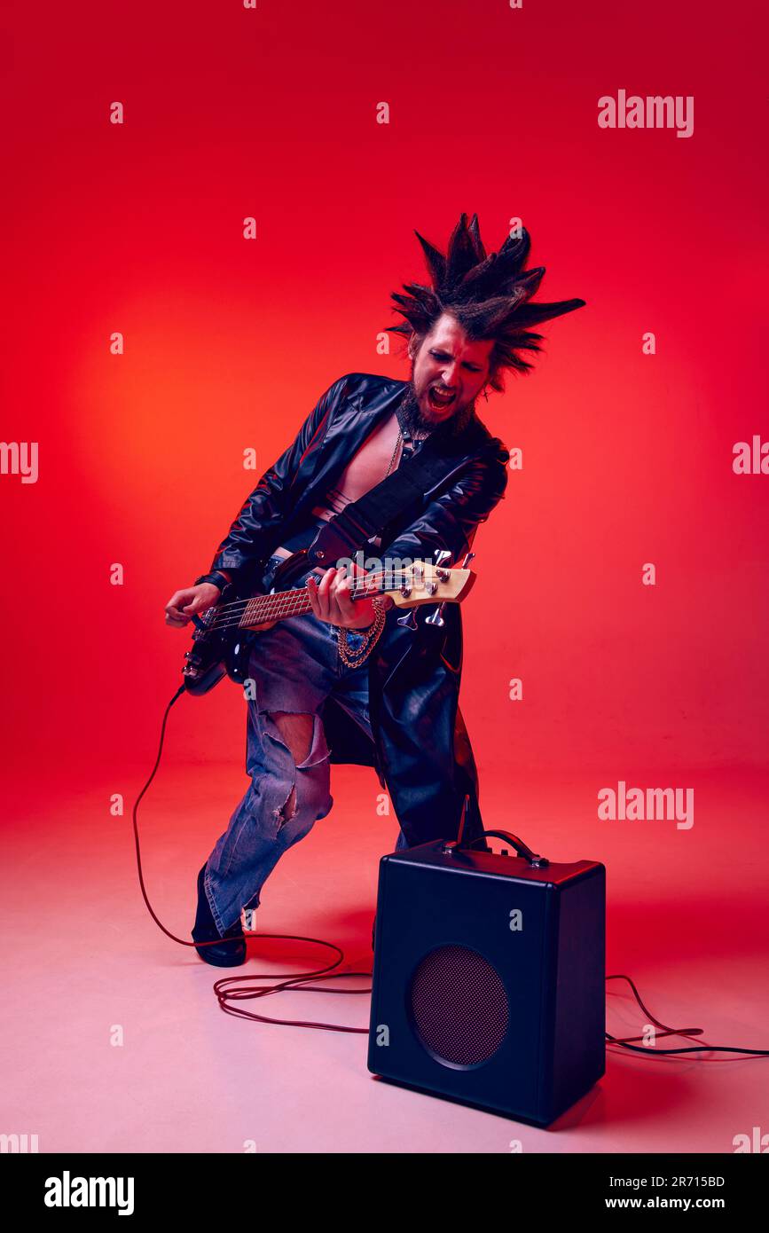 Young man, punk in extraordinary clothes and hairdo playing electric guitar against red studio background in neon light Stock Photo