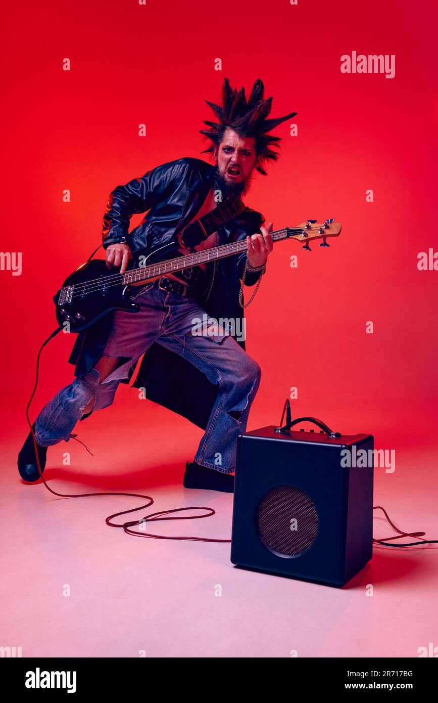 Young emotional man, punk in extraordinary clothes and hairdo playing electric guitar against red studio background in neon light Stock Photo