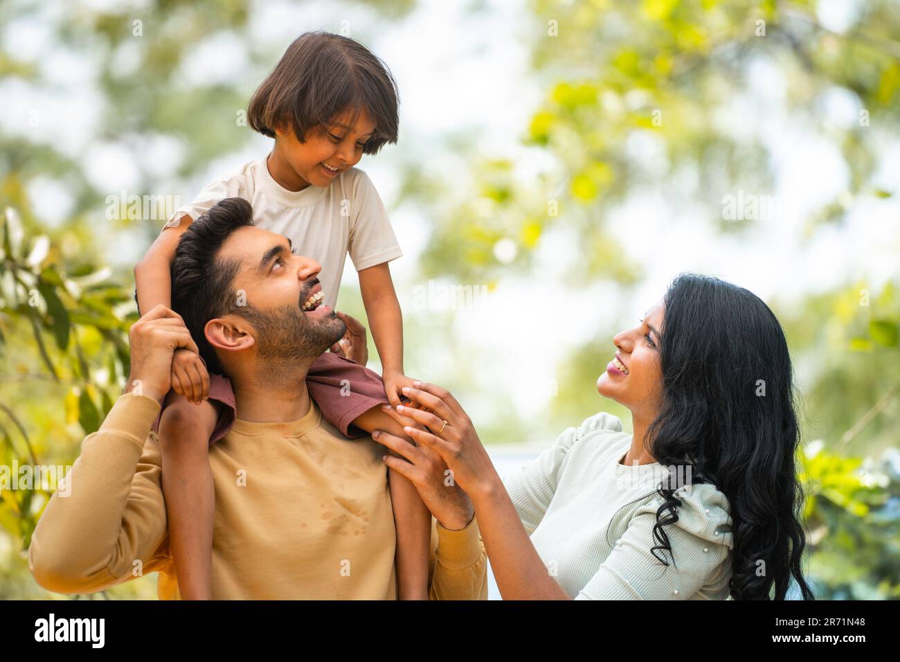 Happy indian parents playing with daughter while kid on father shoulder at outdoor - concept of parenthood, family bonding and weekend holidays Stock Photo