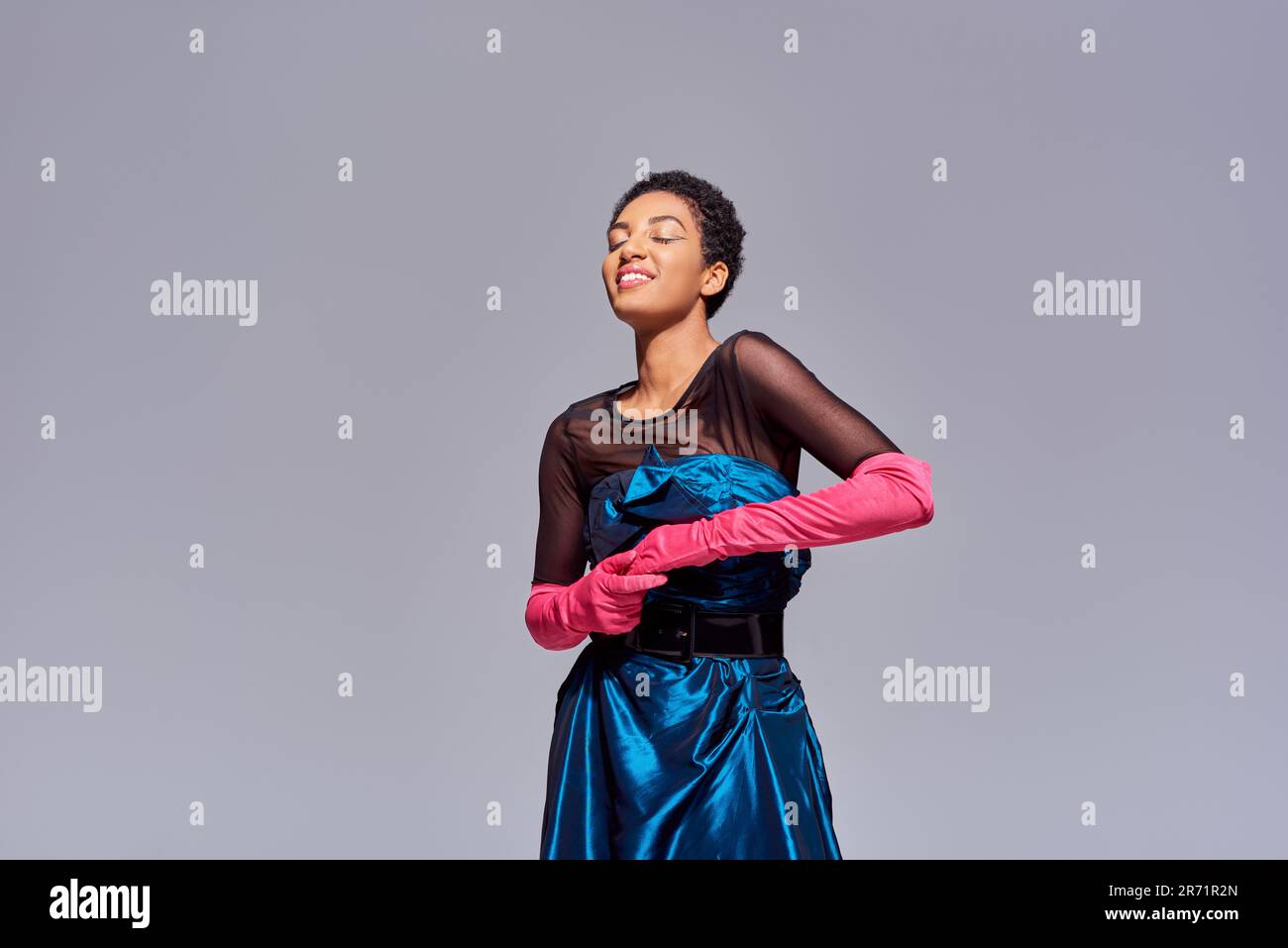Cheerful and stylish young african american woman in pink gloves and  cocktail dress standing with closed eyes isolated on grey, modern  generation z fa Stock Photo - Alamy
