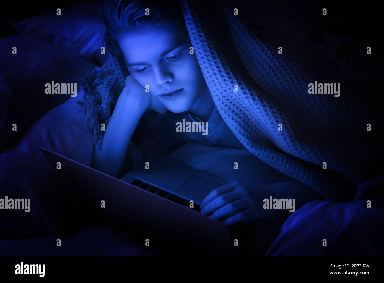 addiction. Teenage boy using laptop under blanket on bed at