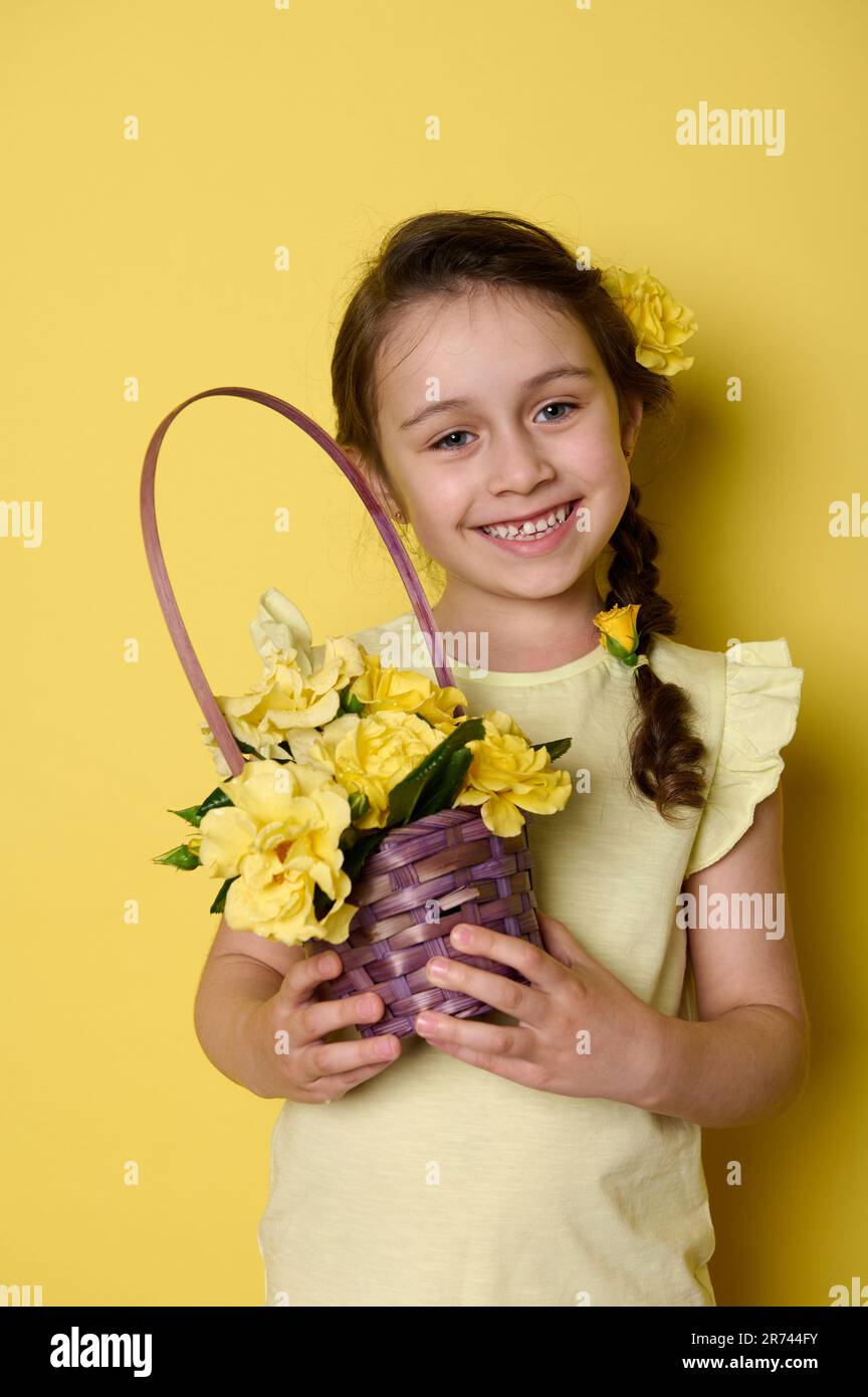 Little girl with flowers in pigtail, dressed in yellow wear, holding purple wicker basket with roses, smiling at camera Stock Photo