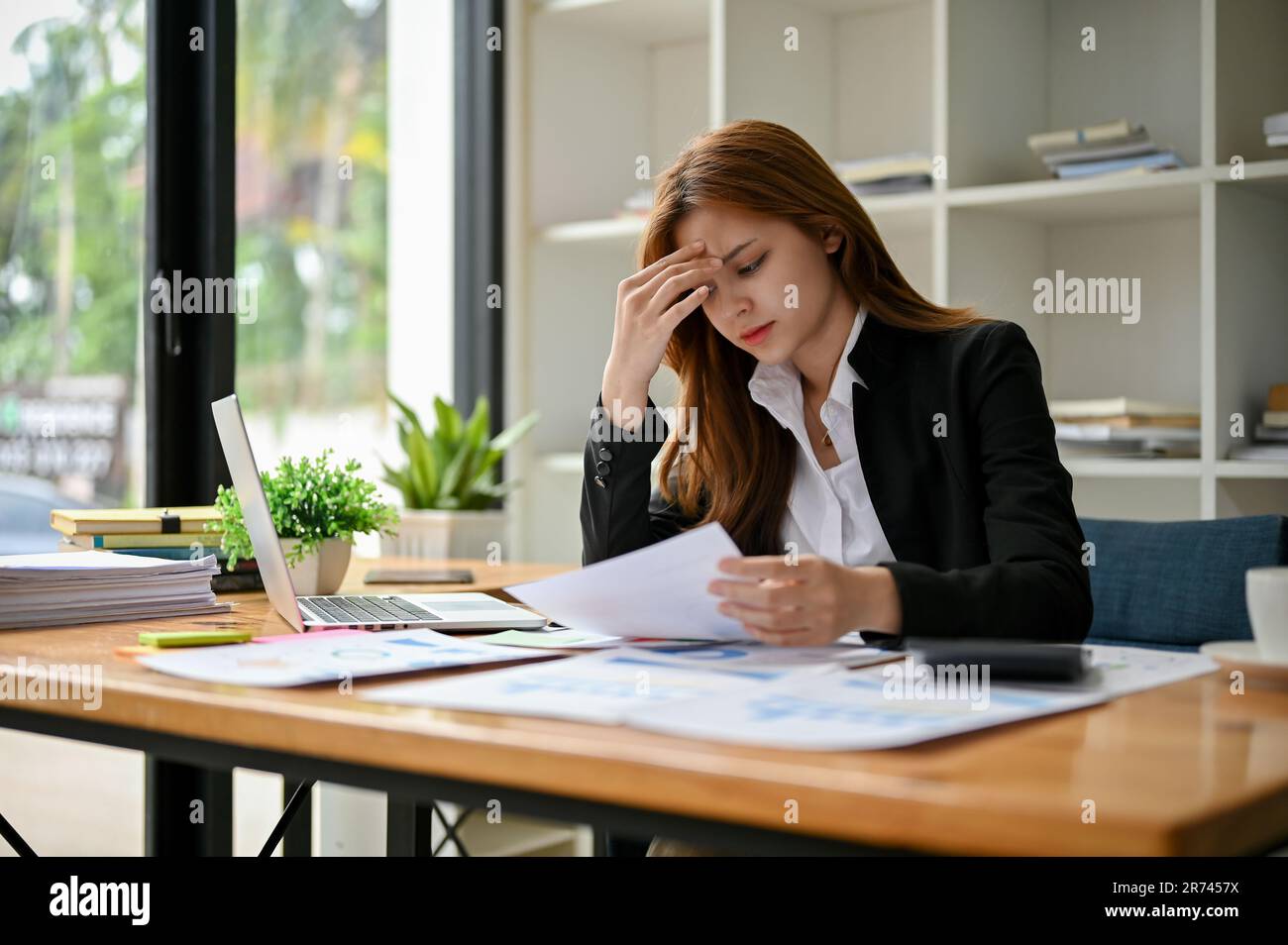 A stressed and serious Asian businesswoman suffers from headaches or eye stains while focusing on reading a business financial report at her desk in t Stock Photo