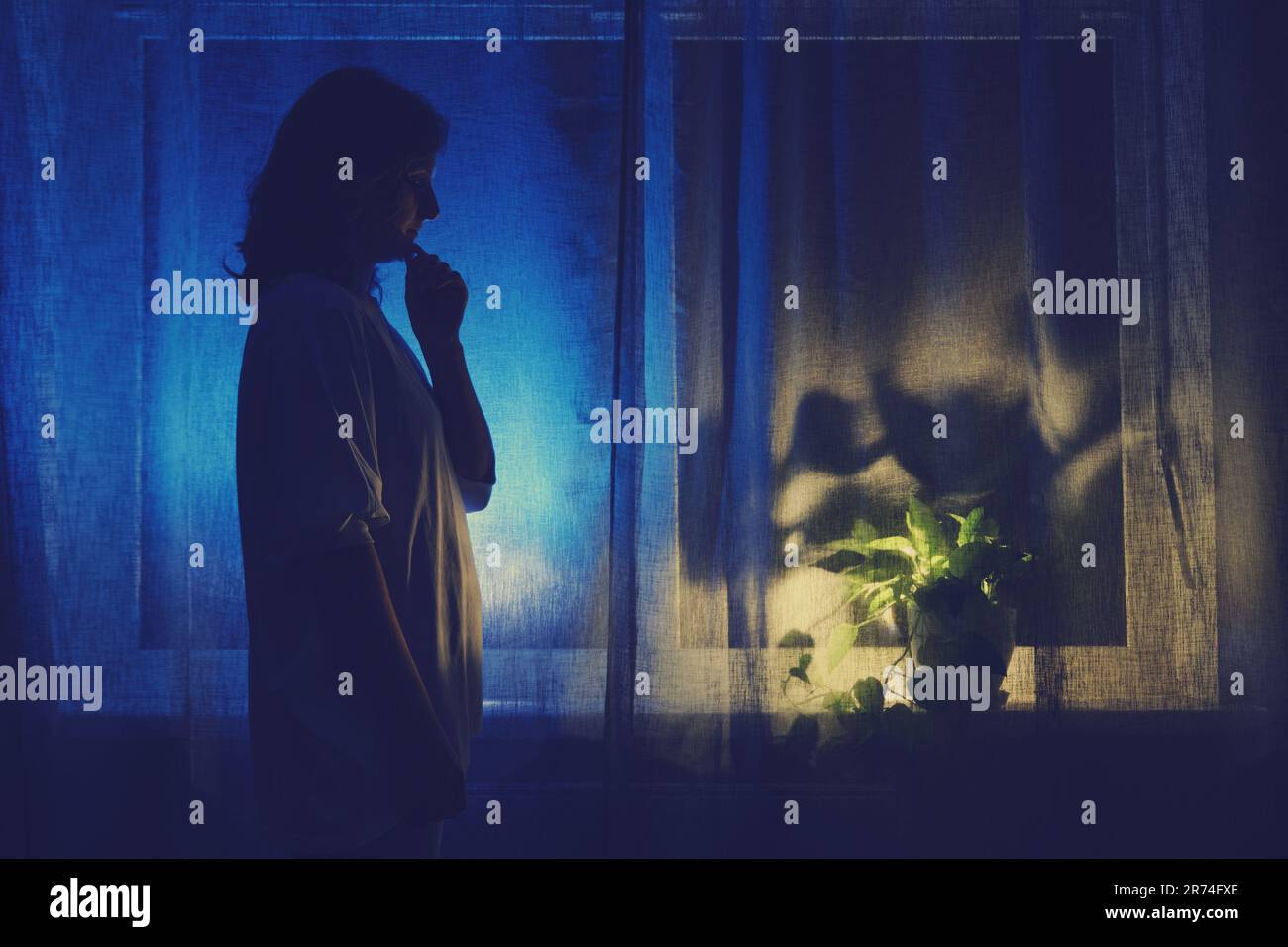 Shadow from a female figure with a thoughtful gesture against the background of a window curtain. A woman is standing in thought at the night dark win Stock Photo