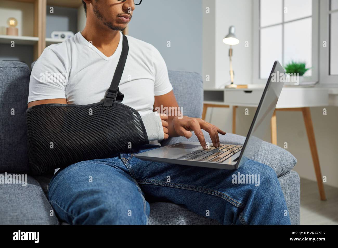 Young man with an arm sling on his broken wrist sitting on the sofa and working on his laptop Stock Photo
