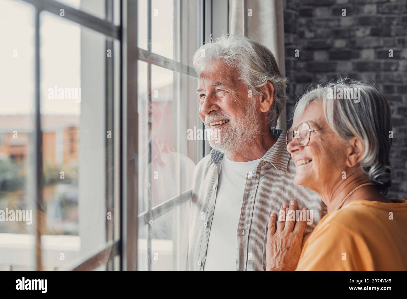 Happy bonding loving middle aged senior retired couple standing near window, looking in distance, recollecting good memories or planning common future, enjoying peaceful moment together at home. Stock Photo
