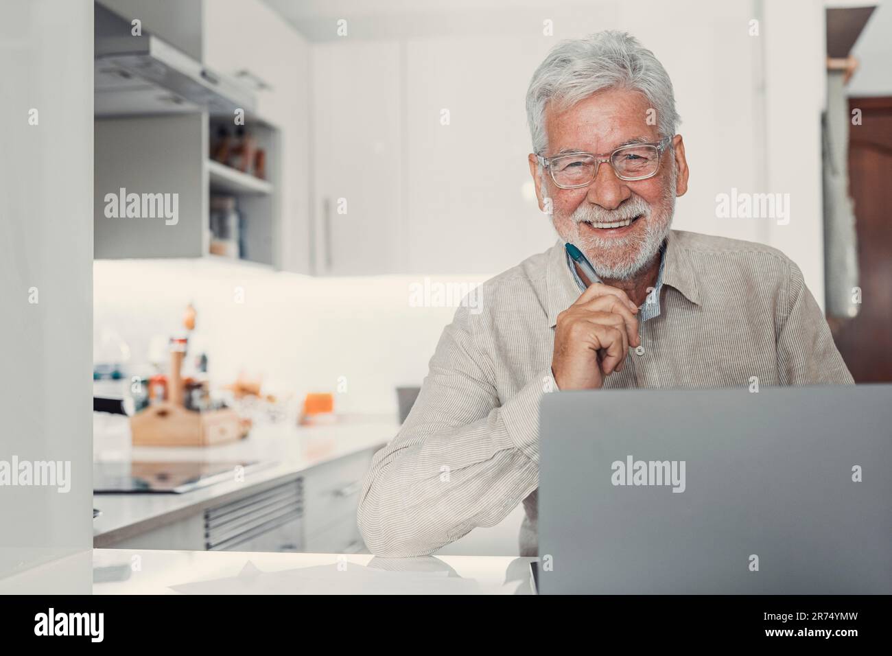 Aged remote worker. Concentrated senior male in glasses work on laptop from home office looking at the camera. Old age man employee freelancer sit at kitchen table by pc typing report online Stock Photo