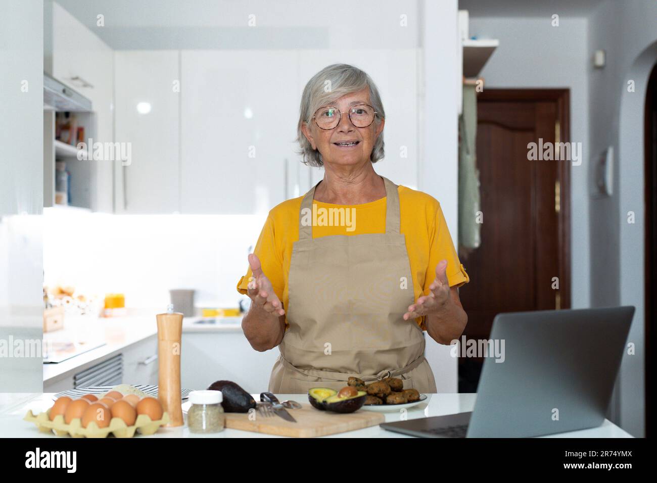 Portrait headshot of old mature woman cooking and preparing meal talking to the camera explaining her recipe to the internet online using laptop. Stock Photo