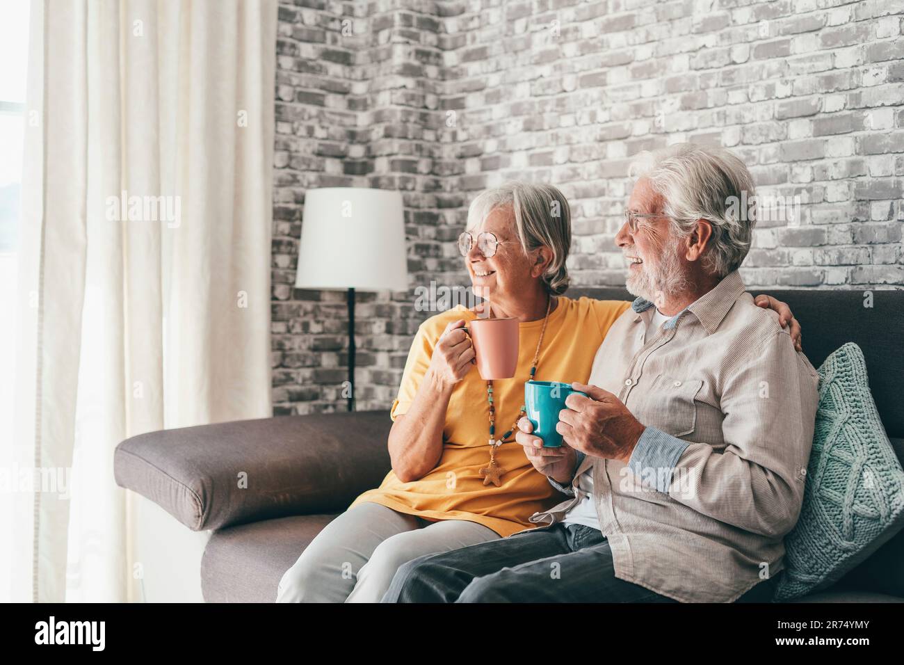 Happy mature husband and wife sit rest on comfortable sofa in living room enjoy tea talking, smiling elderly 60s couple relax on couch at home drink coffee chat speak laugh on leisure weekend Stock Photo