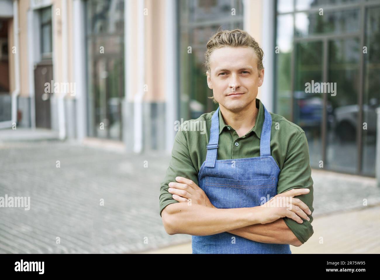 Restaurant or kitchen worker wearing blue apron, standing against backdrop of city. Concept of service and readiness to help. Customer service-oriented nature of the hospitality industry. . High quality photo Stock Photo