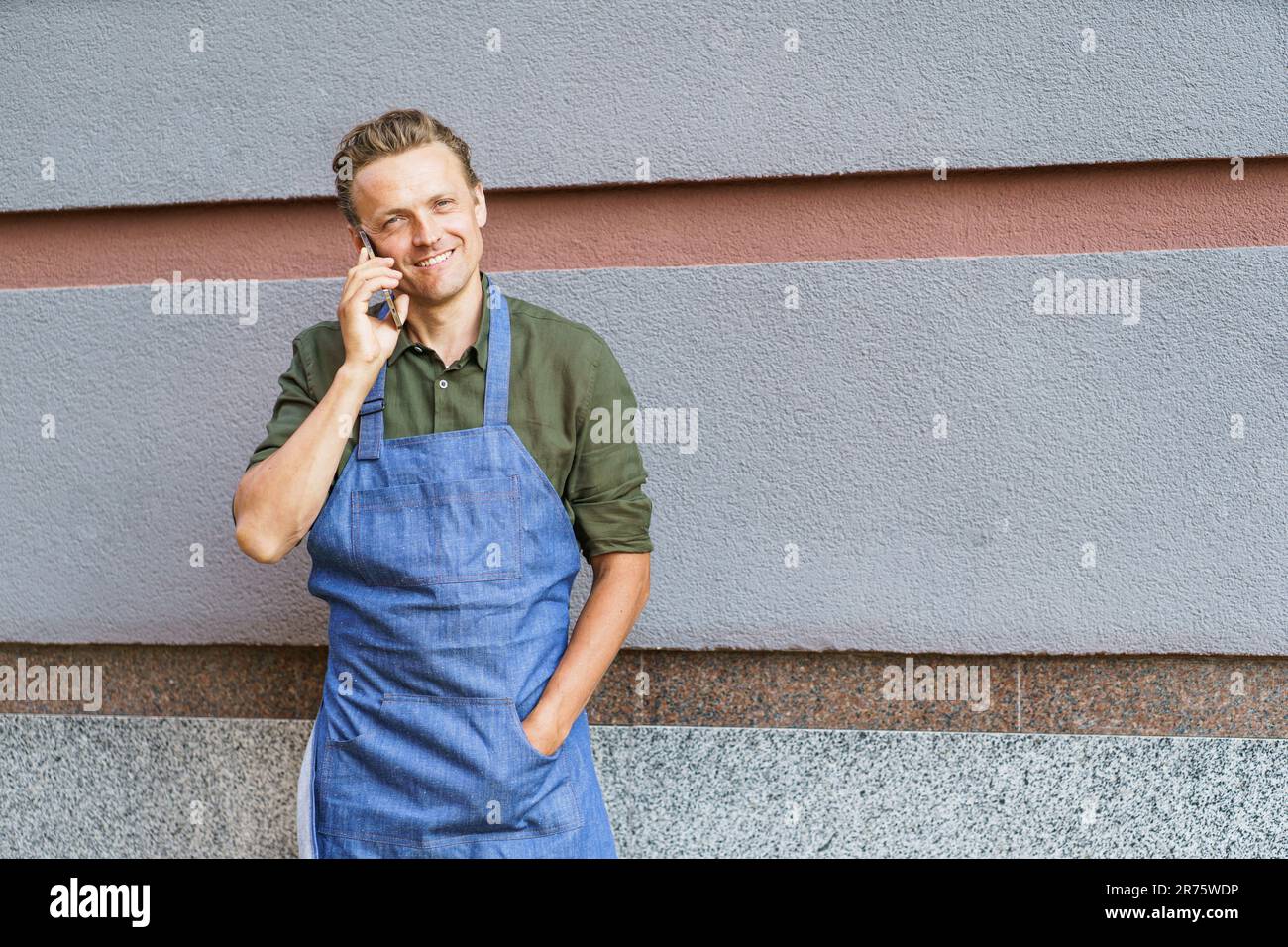 Concept of service readiness and the willingness to assist customers. Worker in blue apron, holding mobile phone, and ready to take order from client against wall background. High quality photo Stock Photo