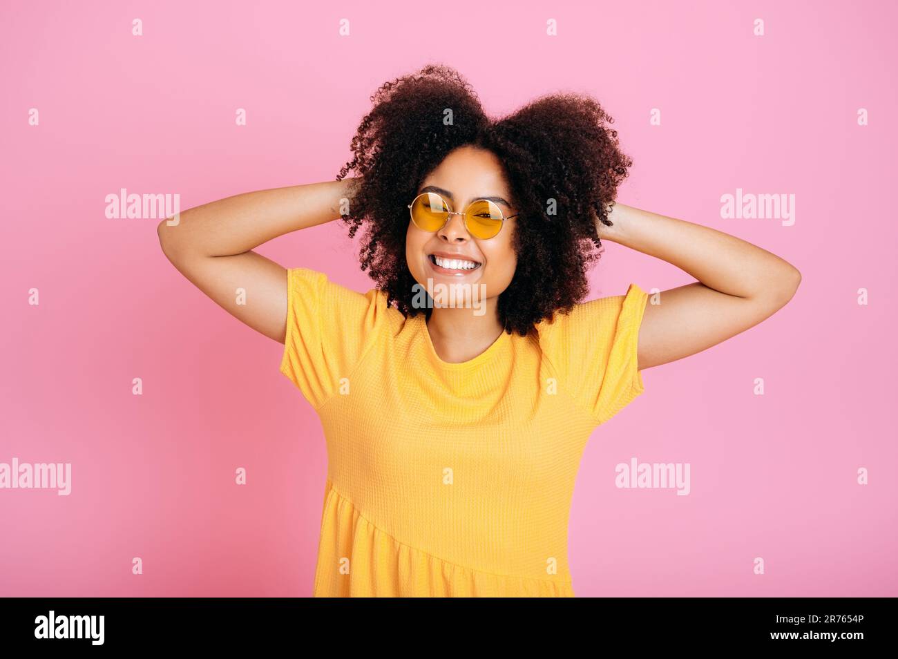 Cheerful excited brazilian or african american curly young woman in trendy orange glasses and sundress, having fun, posing, holding hands on hair, looking at camera, smiling, isolated pink background Stock Photo