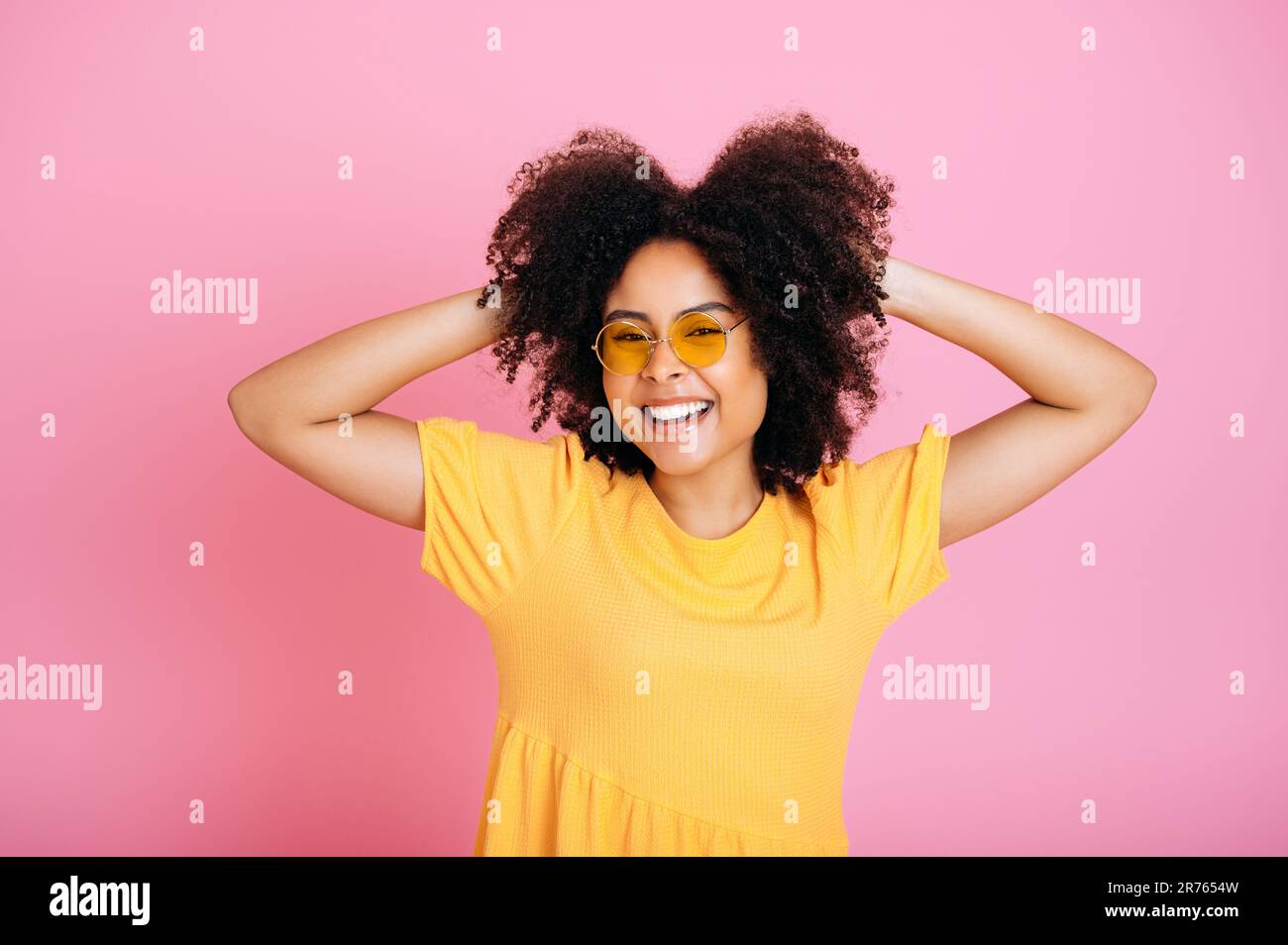 Lovely joyful excited african american or hispanic curly haired woman in trendy orange glasses and sundress, having fun, posing, holding hands on hair, looks at camera, smile, isolated pink background Stock Photo