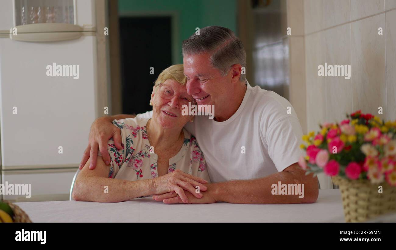 Adult middle-aged son embracing elderly mother while sitting in home kitchen, domestic authentic Heartwarming family moment lifestyle Stock Photo