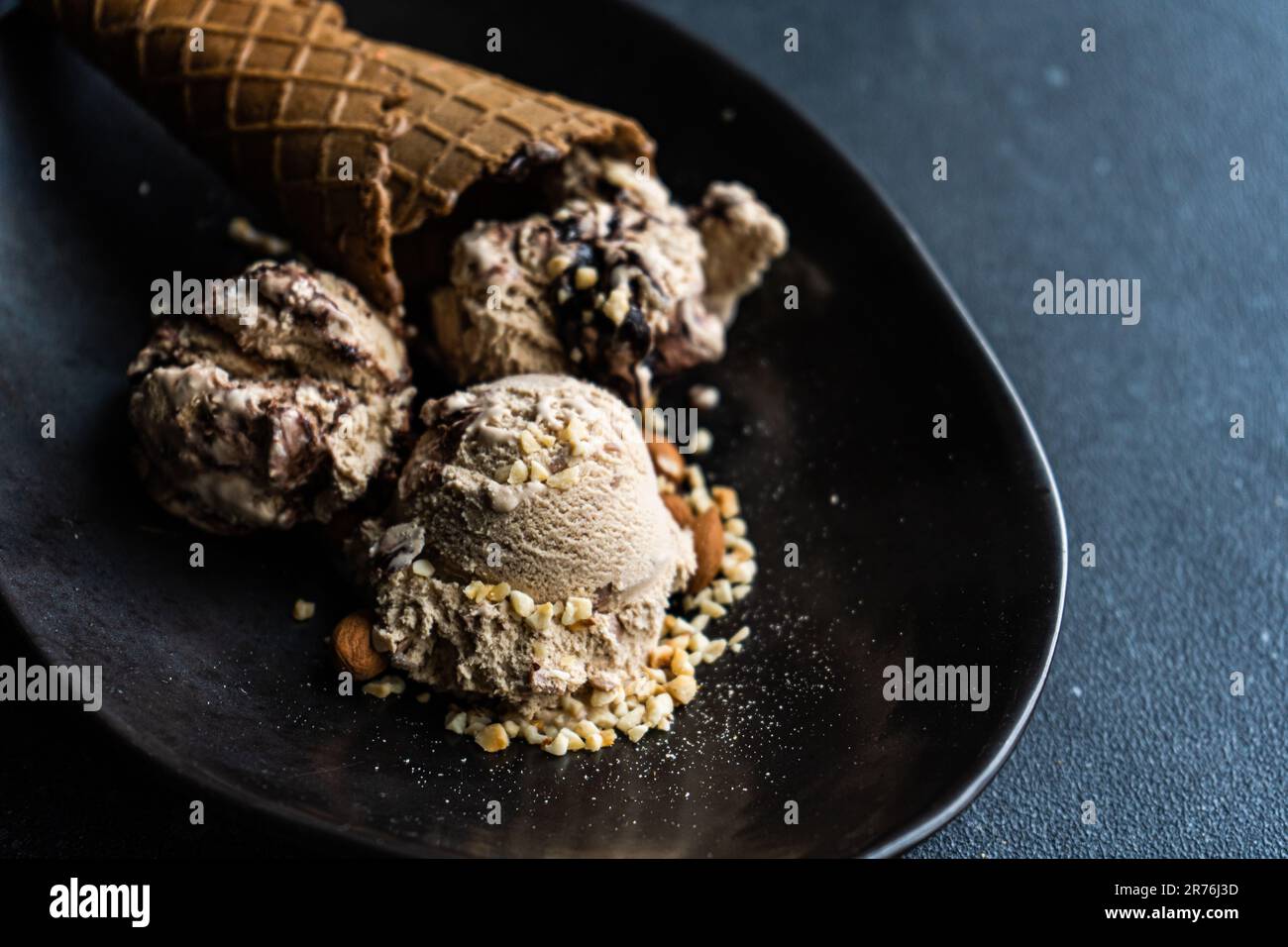 Black ceramic plate with a cone of chocolate ice cream accompanied by two scoops of ice cream with peanuts and walnuts on a dark concrete background Stock Photo