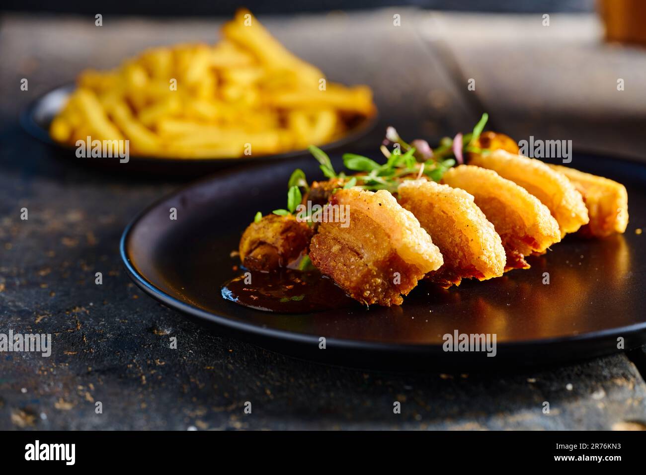 Delicious fried breaded meat garnished with mushrooms and herbs served on black plate near French Fries against blurred background Stock Photo