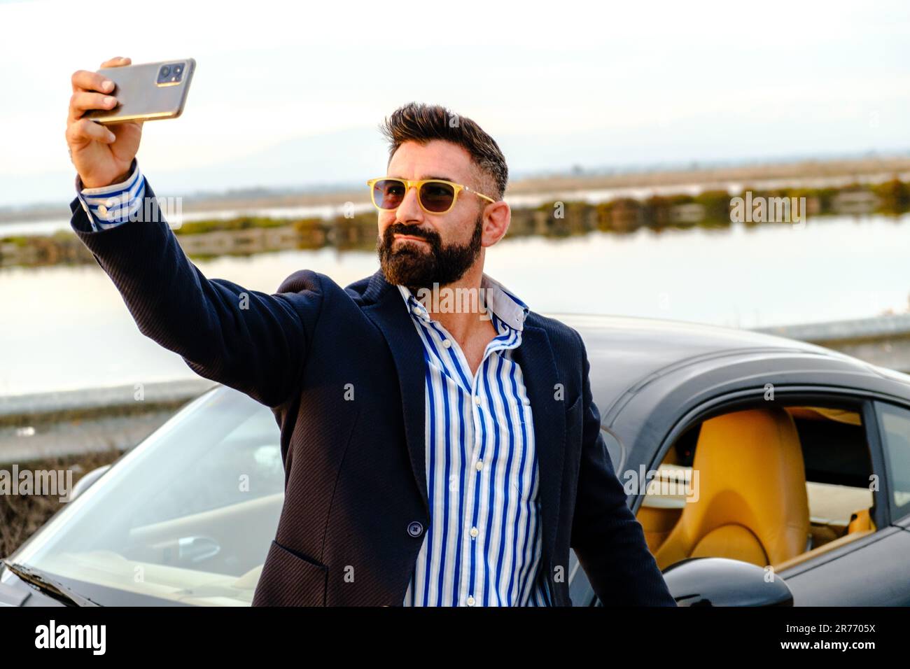 A young male wearing sunglasses taking a selfie while standing next to his sports car Stock Photo
