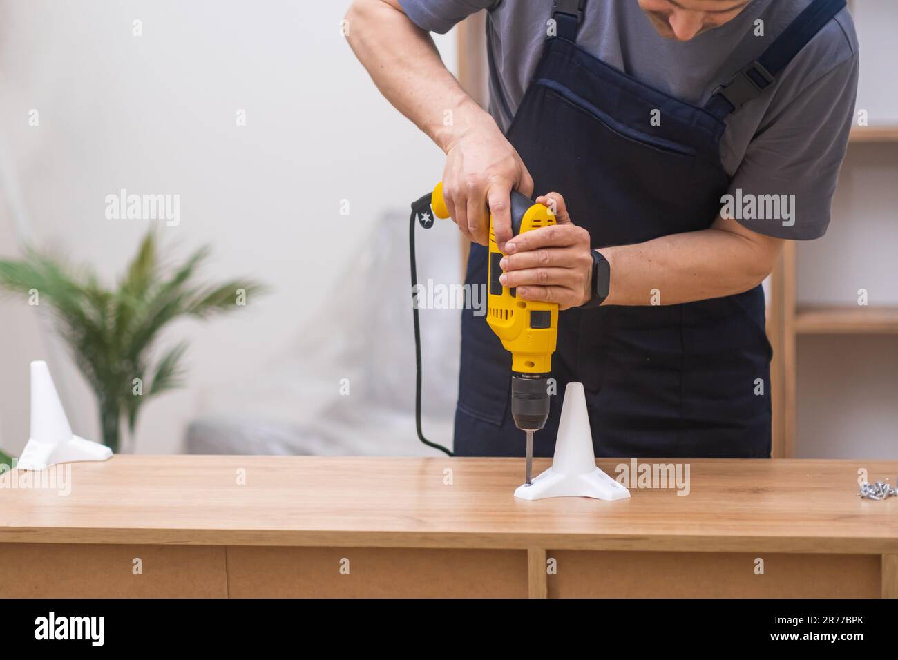 Experienced furniture assembler using an electric screwdriver to attach legs to a wooden rack  Stock Photo