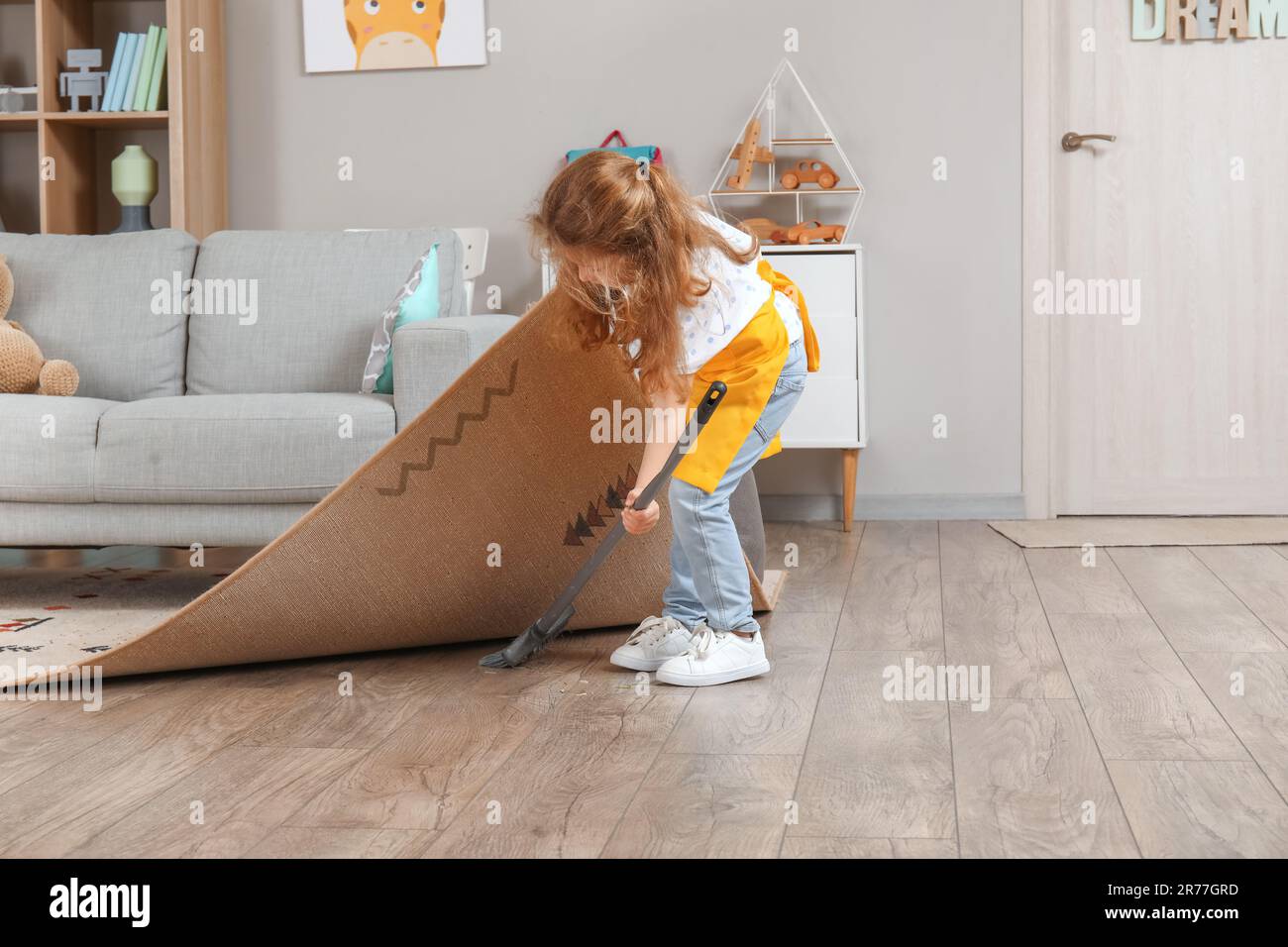 Cute little girl sweeping floor under carpet with broom at home Stock