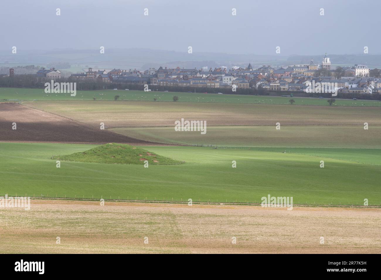 An ancient tumulus burial mound stands in a field below Maiden Castle in Dorset, with Poundbury new town in the distance. Stock Photo