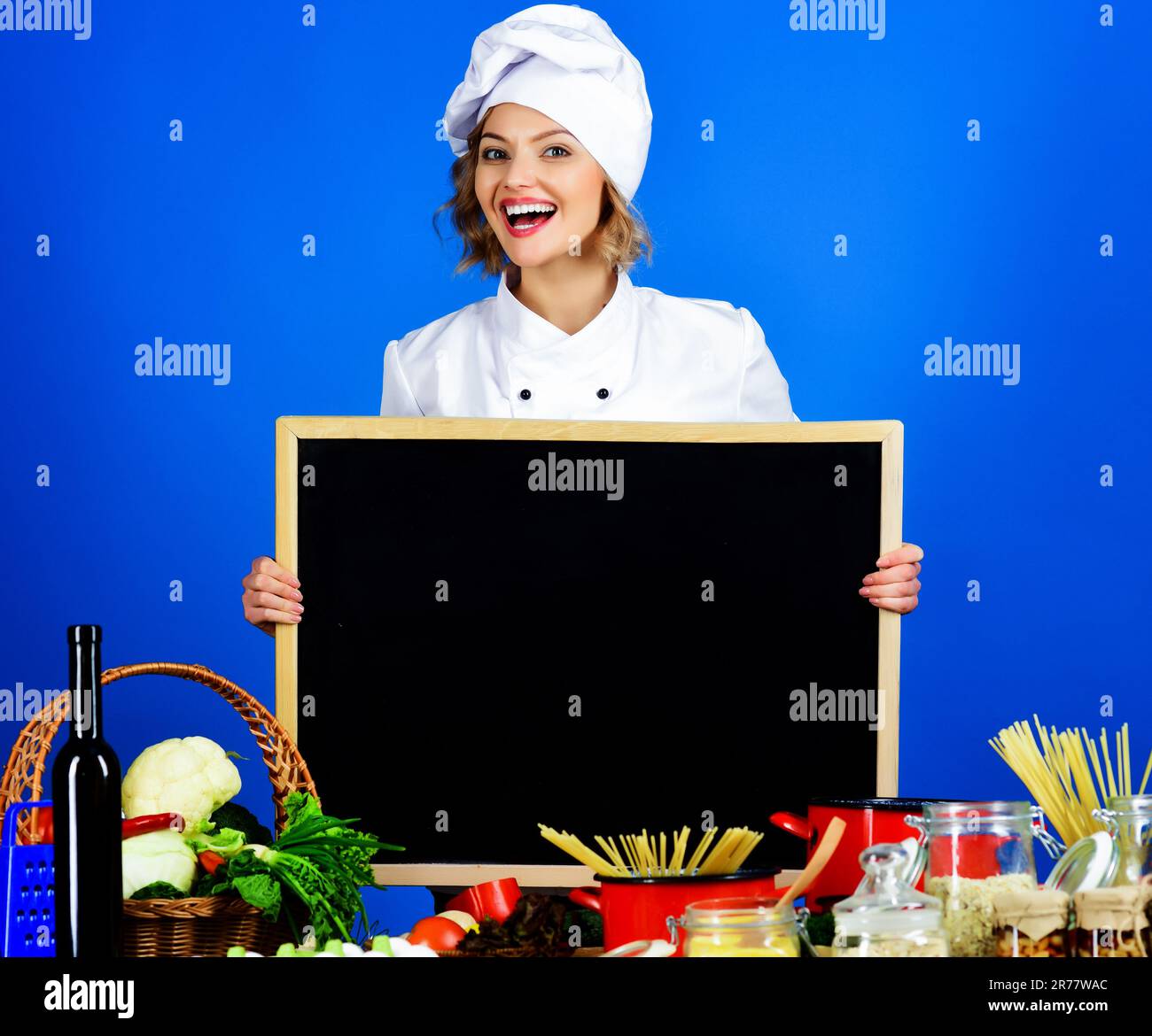 Smiling female chef with blank blackboard. Food preparation. Healthy menu. Cook in chef uniform with empty billboard. Copy space for advertise menu in Stock Photo