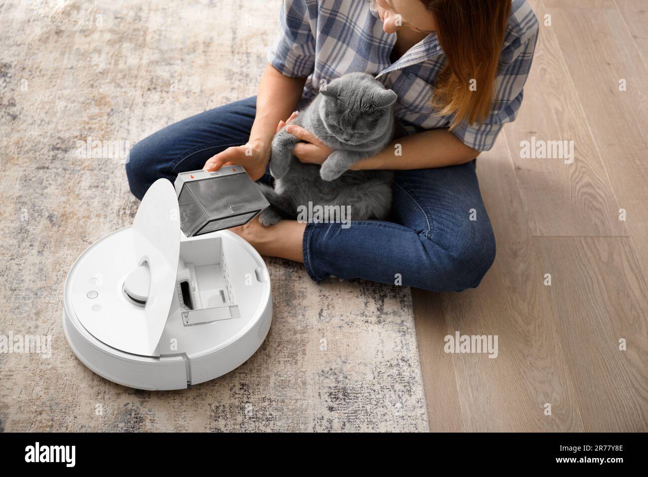 The container of the vacuum cleaner robot is in female hands, the dust collector of the robot vacuum cleaner is full of wool, the cat sits nearby Stock Photo