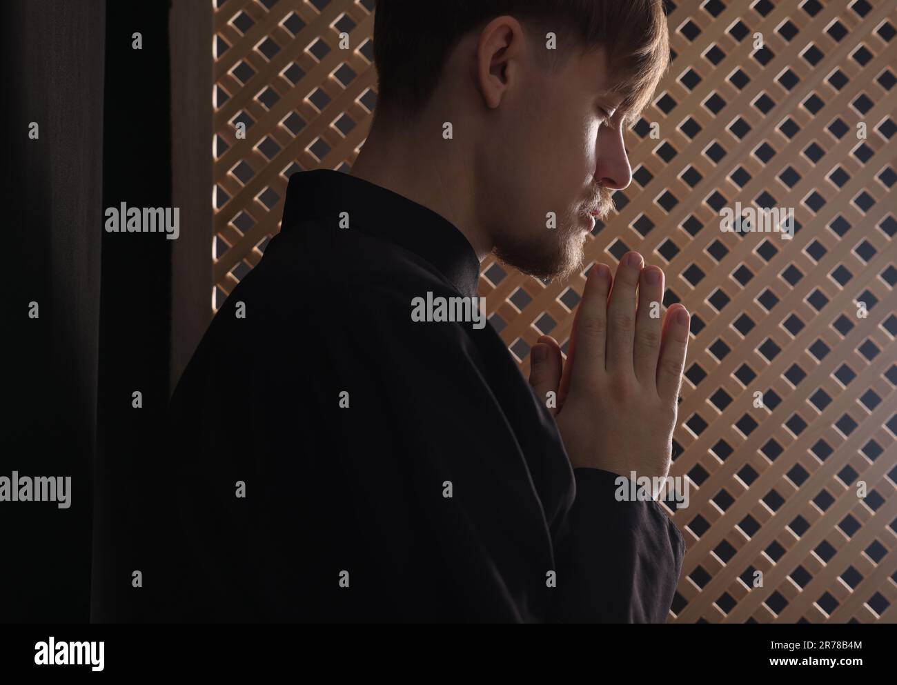 Catholic priest praying near wooden window in confessional booth Stock Photo