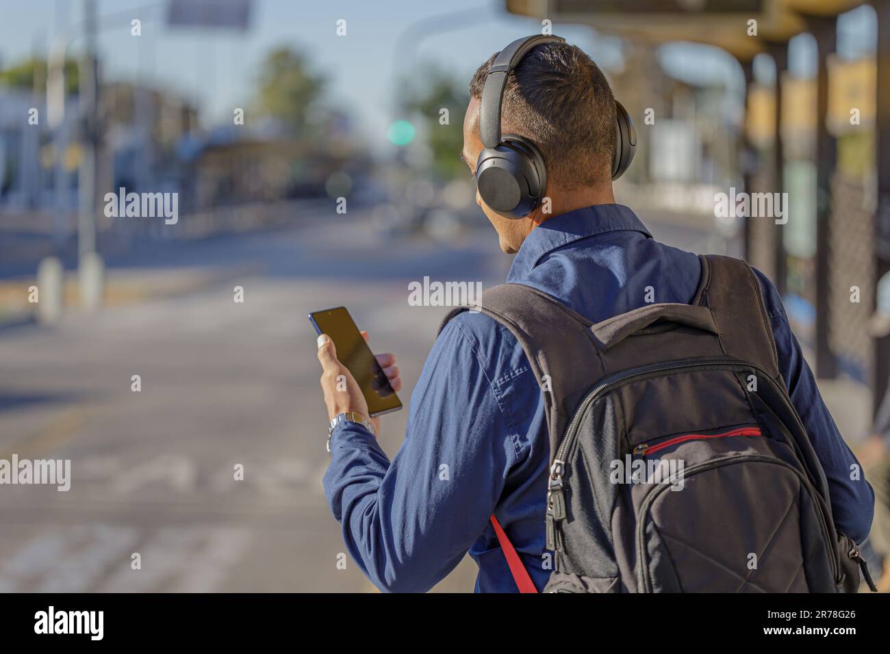Young latin man with headphones looking at his mobile phone at the bus stop seen from behind with copy space. Stock Photo