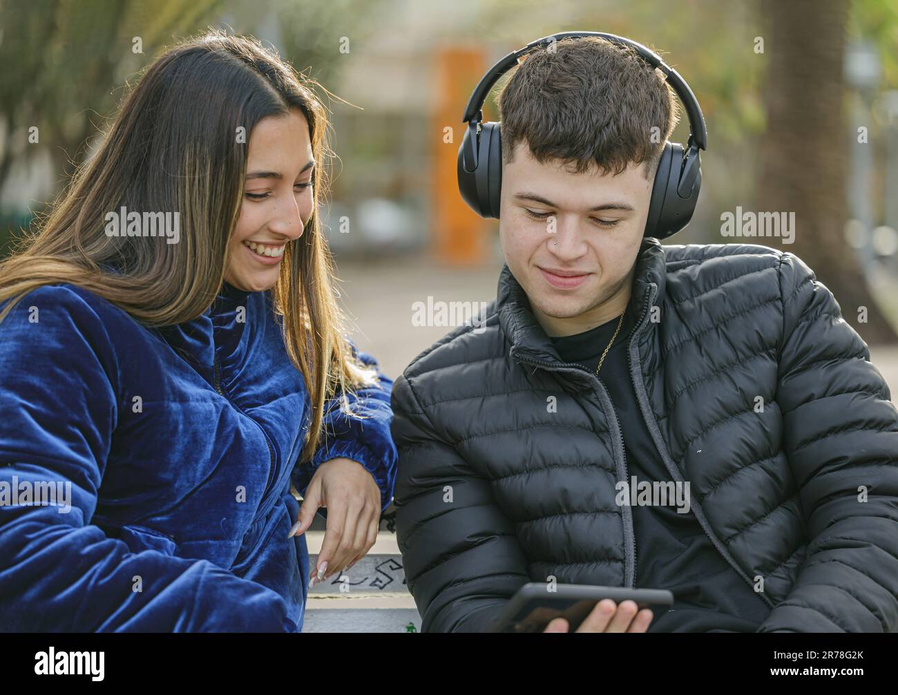 Young couple sitting on the bench in a square with mobile phone and headphones. Stock Photo