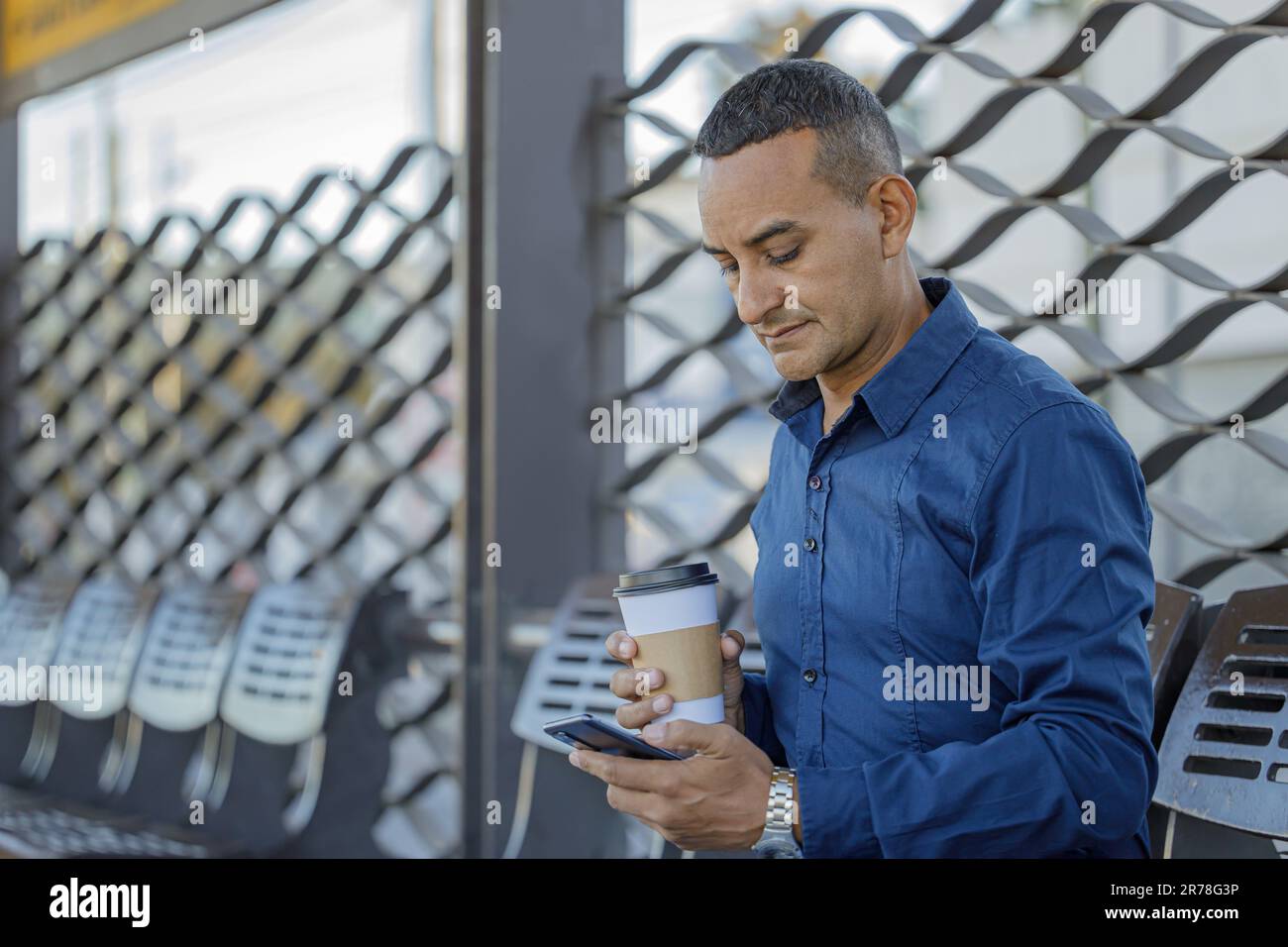 Young latin man looking at his mobile phone at a bus stop with copy space. Stock Photo
