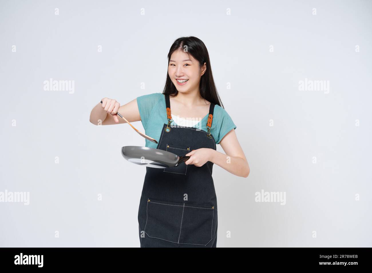 Young woman wearing kitchen apron cooking and holding pan and spatula isolated on white background. Stock Photo