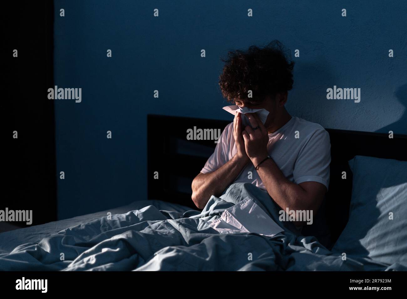 Handsome young man suffering cold and flu trying to sleep at night, guy sneezing into a tissue