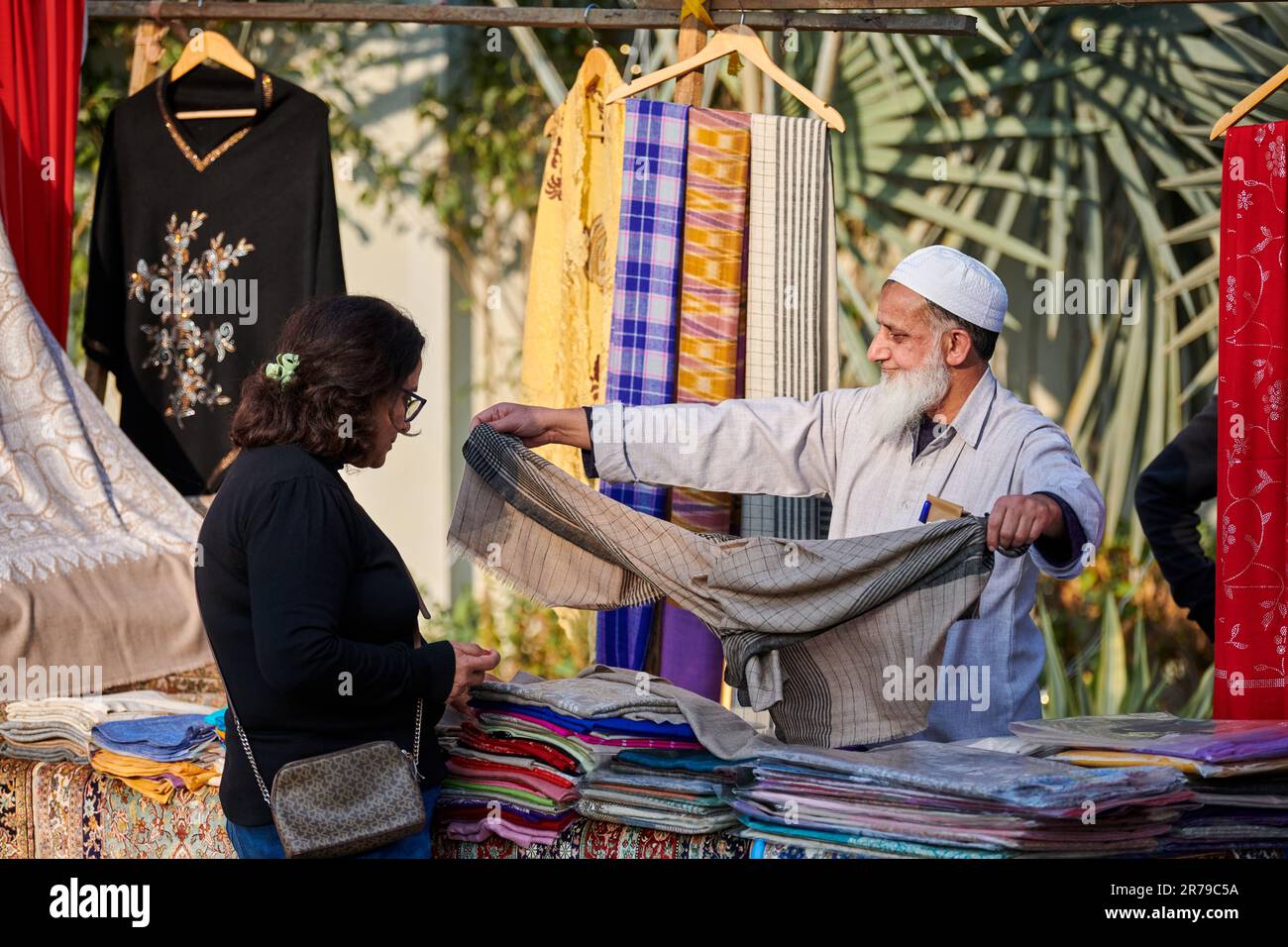 New Delhi, India - 10.12.2022 - Old seller of textile and carpet shows goods to woman at outdoor fair, bearded muslim salesman in taqiyah cap shows ex Stock Photo