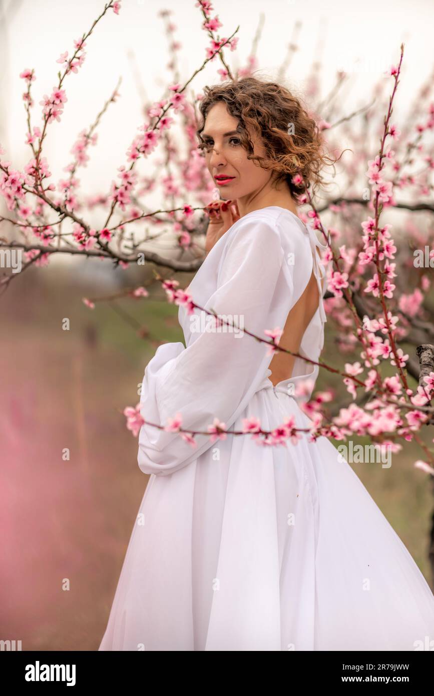 Woman peach blossom. Happy curly woman in white dress walking in the garden of blossoming peach trees in spring Stock Photo