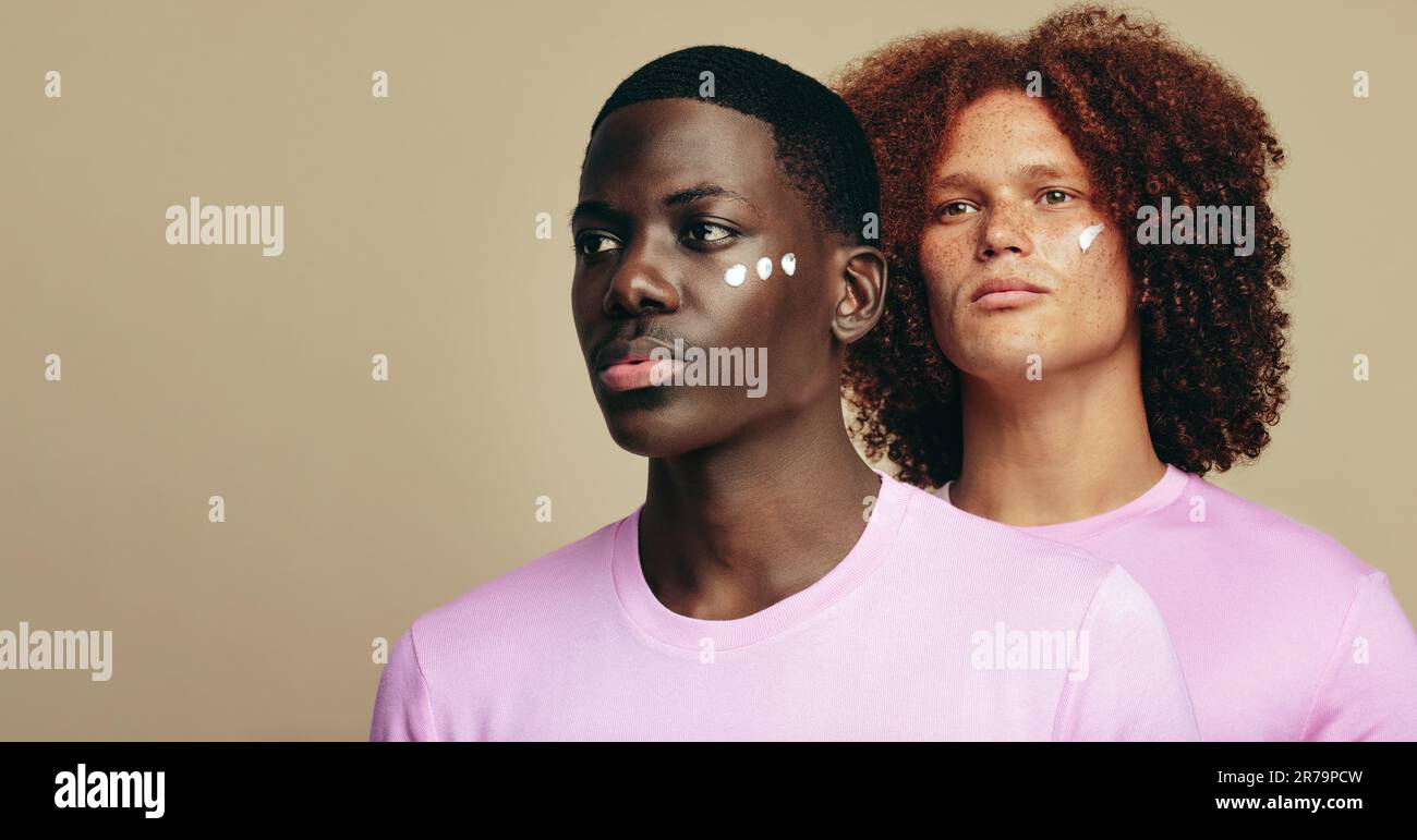 Men with different skin types stand side by side in a studio, moisturizing their faces with a beauty cream. Two diverse young men using a cosmetic pro Stock Photo