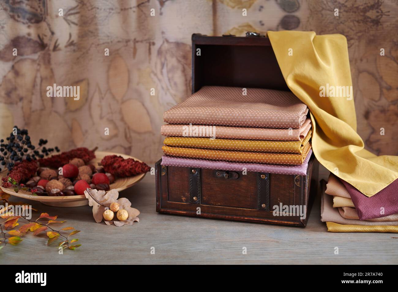 Wooden chest with stack of hand-dyed fabrics, wooden plate with autumn leaves, nuts and berries on an eco-print background Stock Photo