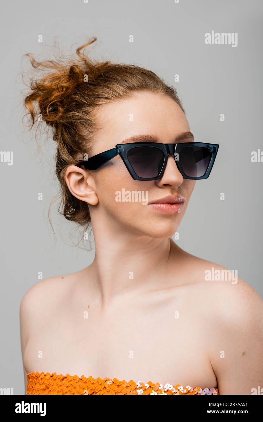 Portrait of young red haired and freckled woman in sunglasses and top with sequins looking away while standing isolated on grey background, trendy sun Stock Photo