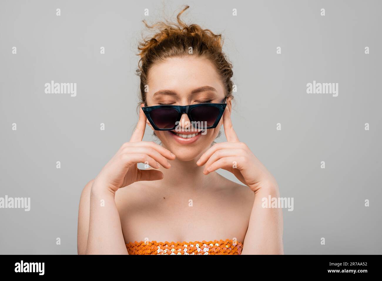 Cheerful young red haired woman with natural makeup in sunglasses and orange top with sequins closing eyes while standing isolated on grey background, Stock Photo