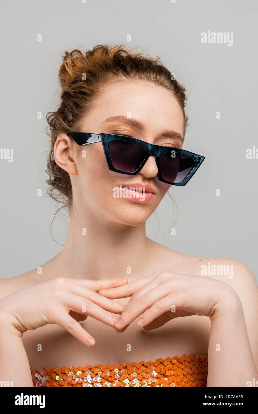 Portrait of young redhead woman with freckles wearing top with sequins and stylish sunglasses while standing isolated on grey background, trendy sun p Stock Photo