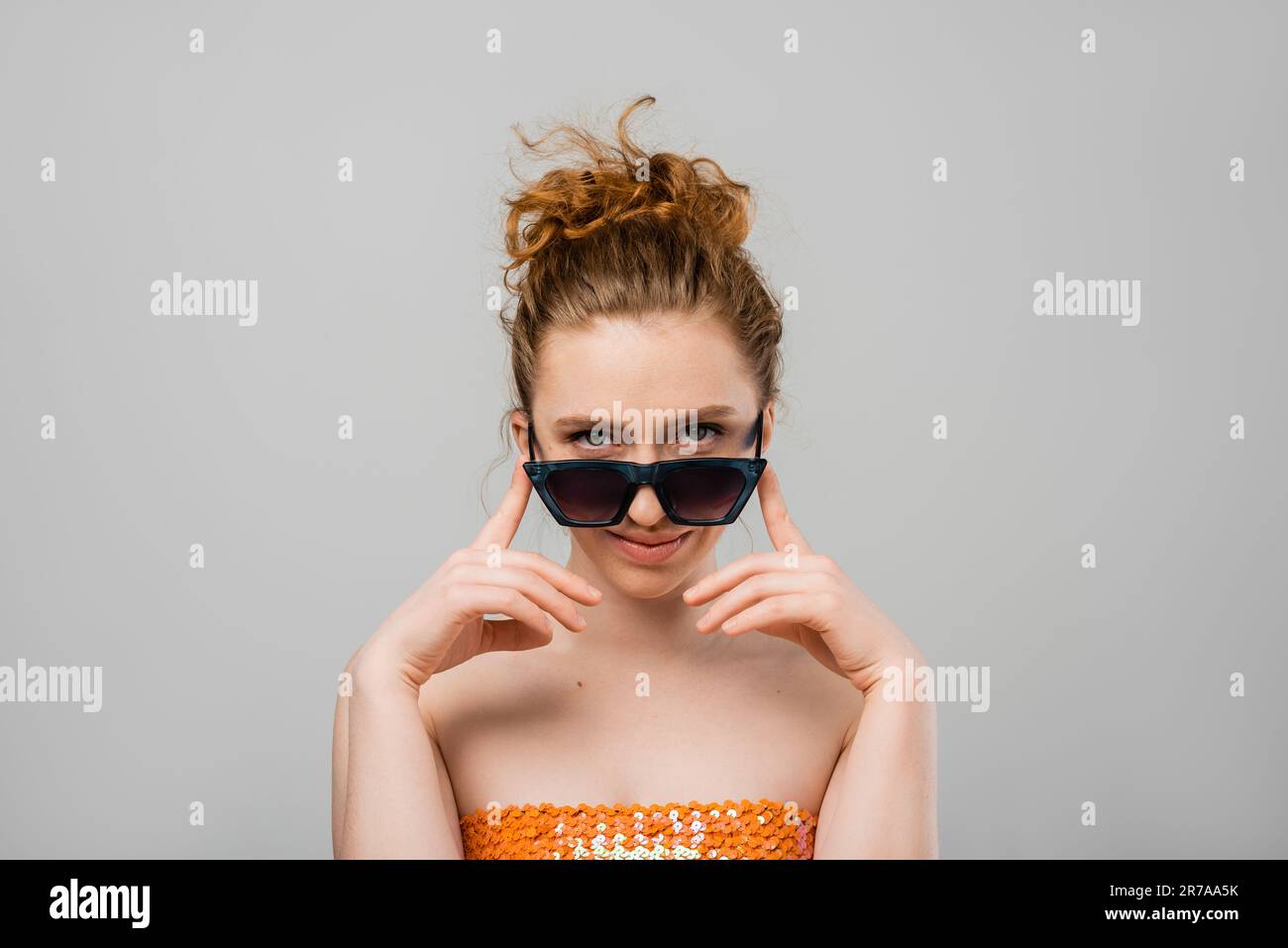Portrait of stylish young red haired woman with natural makeup in top with sequins and sunglasses looking at camera isolated on grey background, trend Stock Photo