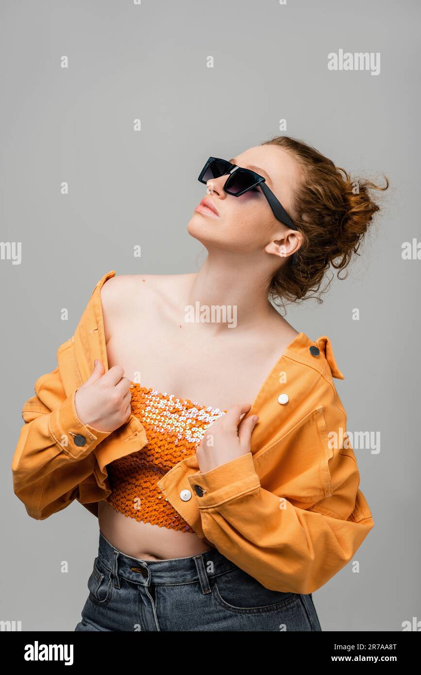 Stylish young redhead woman in sunglasses and top with sequins touching orange denim jacket and standing and posing isolated on grey background, trend Stock Photo