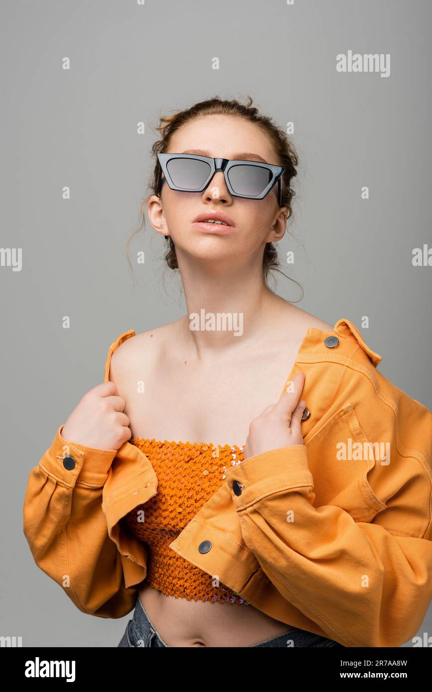 Fashionable red haired model in sunglasses and orange top with sequins touching denim jacket while standing and posing isolated on grey background, tr Stock Photo