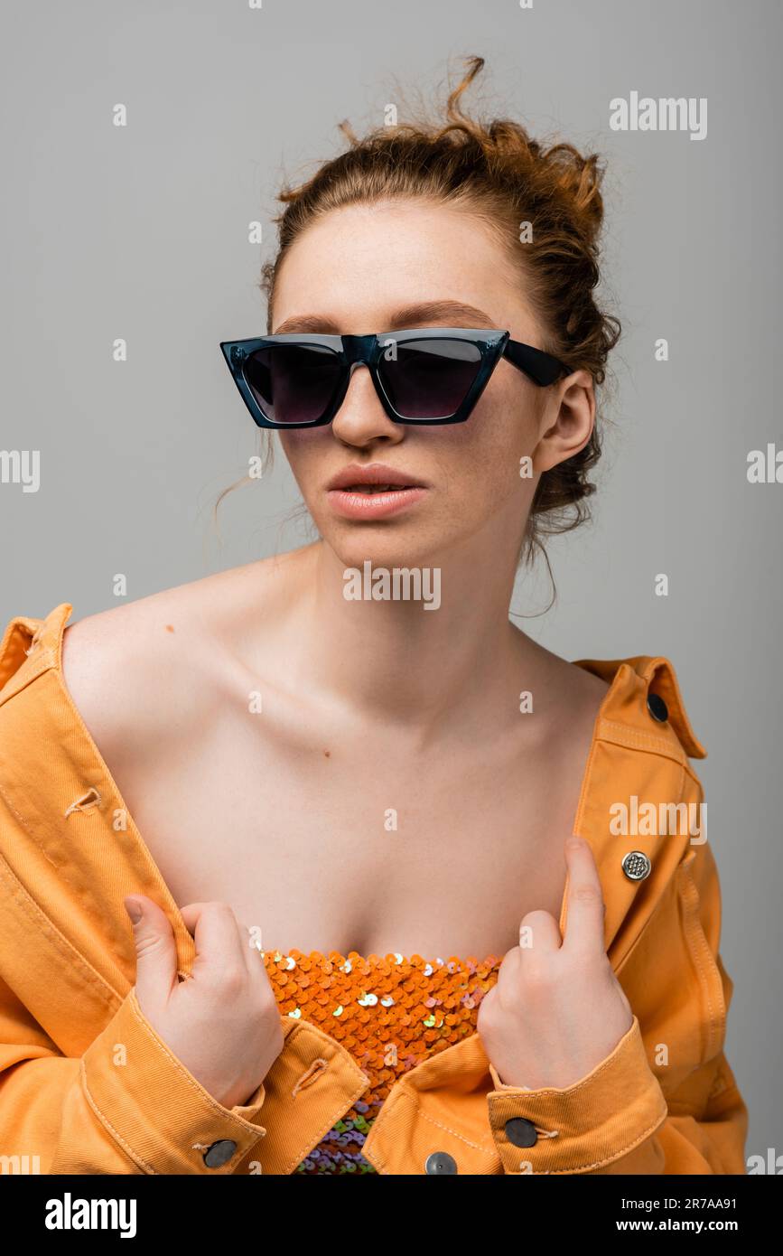 Young and redhead woman in sunglasses and top with orange sequins touching orange denim jacket and posing isolated on grey background, trendy sun prot Stock Photo