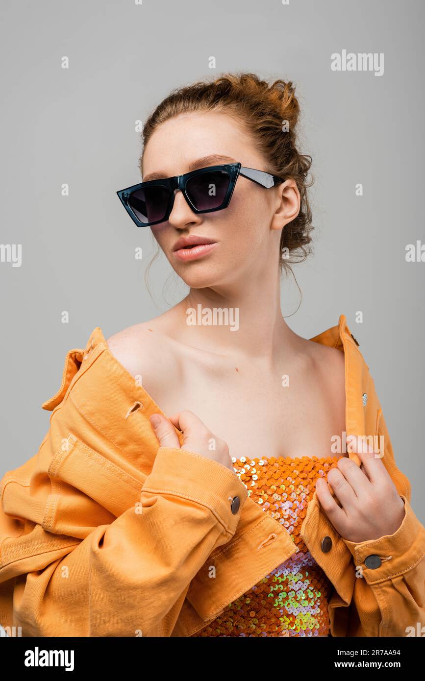 Fashionable redhead woman with natural makeup in sunglasses and top with sequins touching orange jacket while standing isolated on grey background, tr Stock Photo