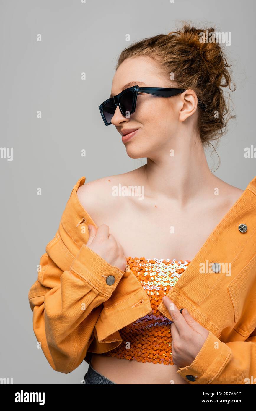 Joyful redhead woman with natural makeup in sunglasses, orange denim jacket and top with sequins posing and standing isolated on grey background, tren Stock Photo